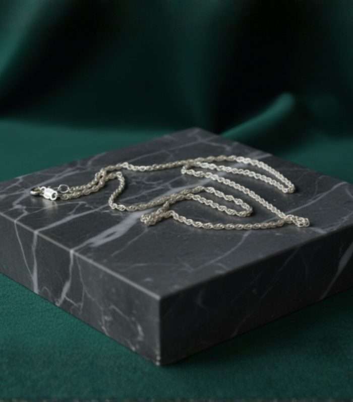 Cadena de plata con diseño elegante, sobre base de mármol oscuro con fondo verde.